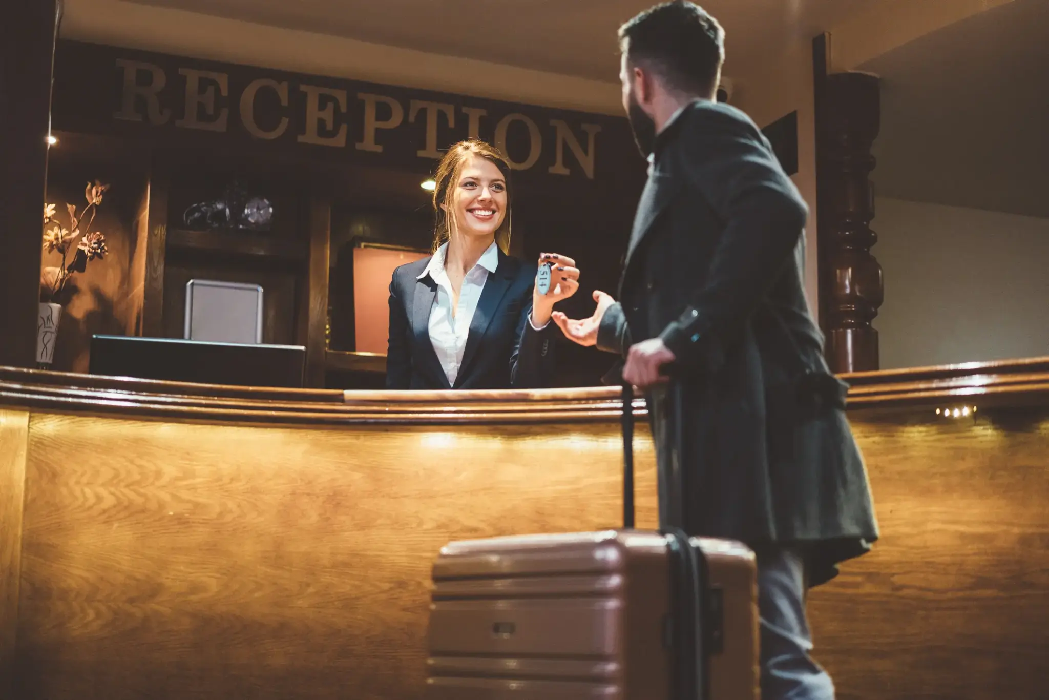 Hotelversicherungen | Rezeptionistin übergibt Hotelschlüssel an ankommenden Gast mit Koffer
