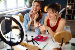 Zwei junge Influencerinnen sitzen lachend vor einer Kamera und nehmen ein Schminkvideo auf, umgeben von Make-up-Produkten und Ringlicht