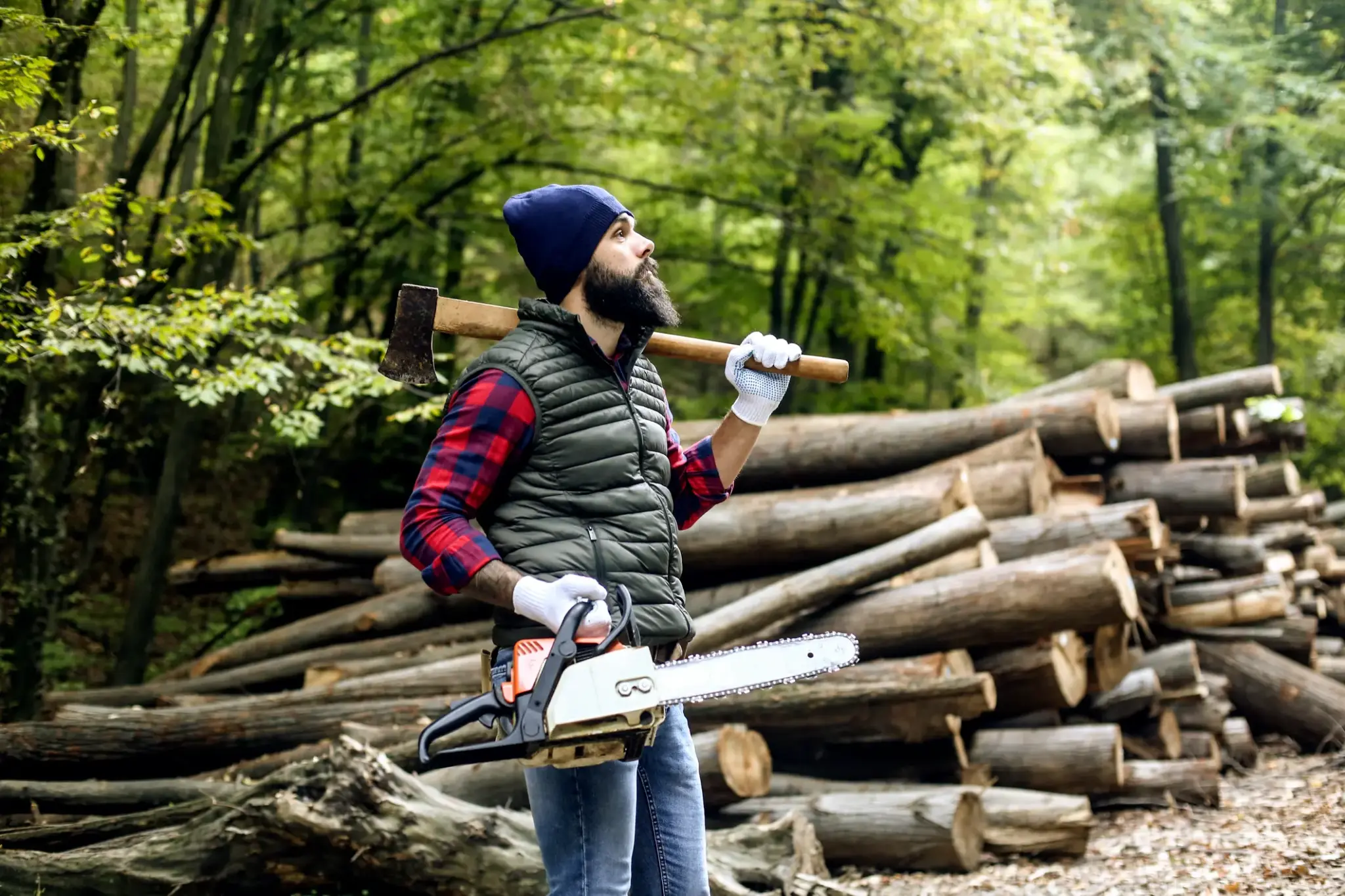 Forstwirt mit Axt und Motorsäge steht vor gestapeltem Holz im Wald und blickt zur Seite während einer Arbeitspause