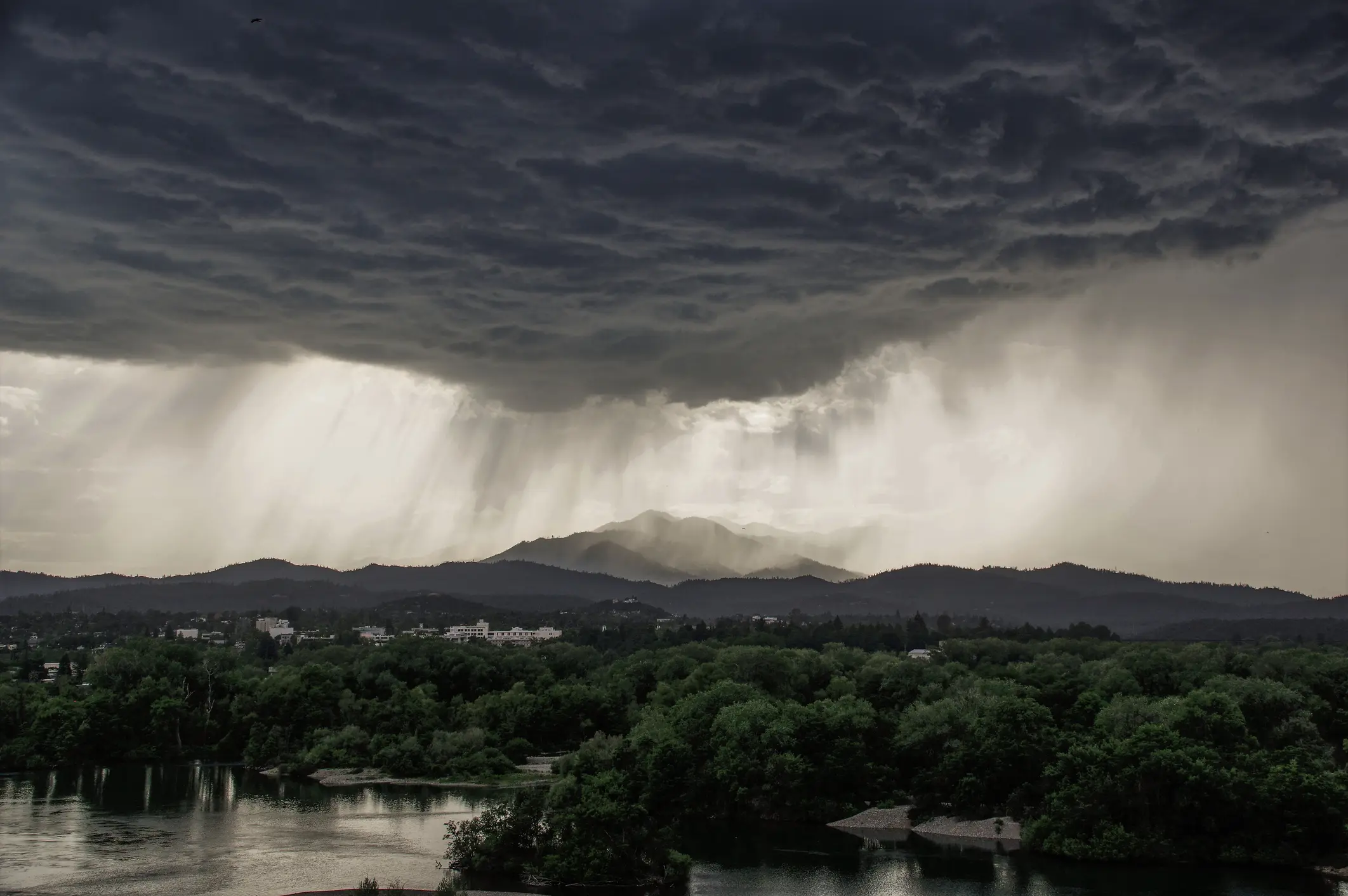 Gewitterfront mit Starkregen und dunklen Wolken über bewaldeter Landschaft und Stadtgebiet