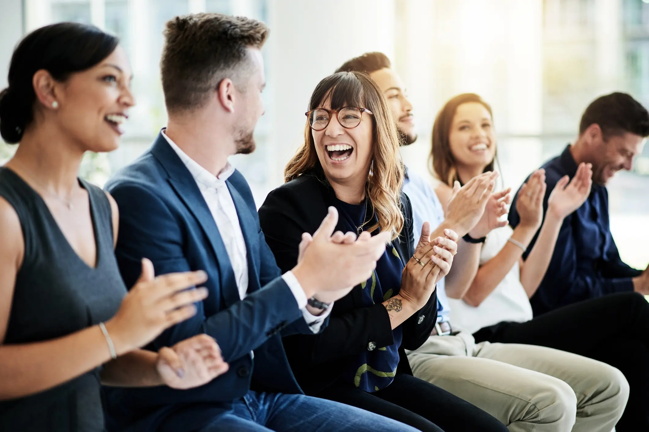 Junge Berufstätige sitzen nebeneinander im Büro, lachen gemeinsam und applaudieren in einer entspannten Teamatmosphäre
