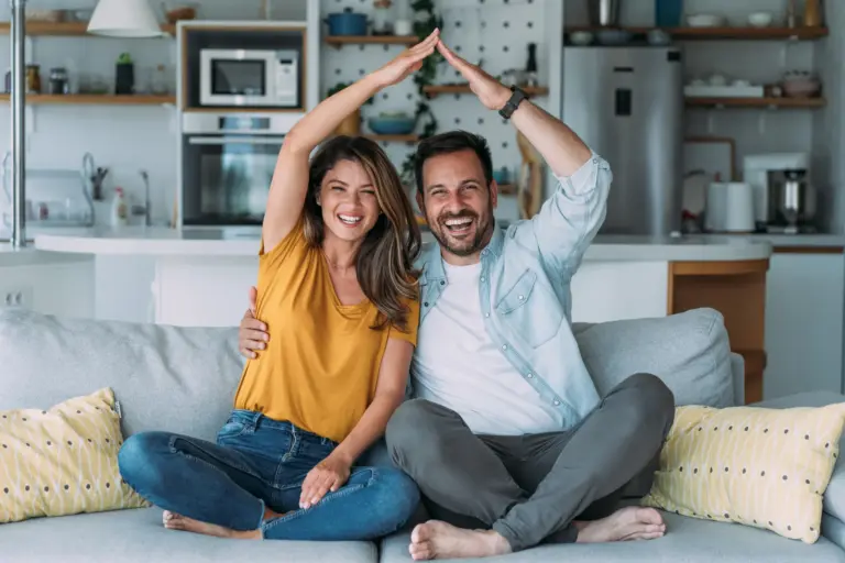 Lachendes Paar sitzt auf dem Sofa und formt mit den Händen ein Hausdach – Symbol für Sicherheit rund um Haus und Wohnen