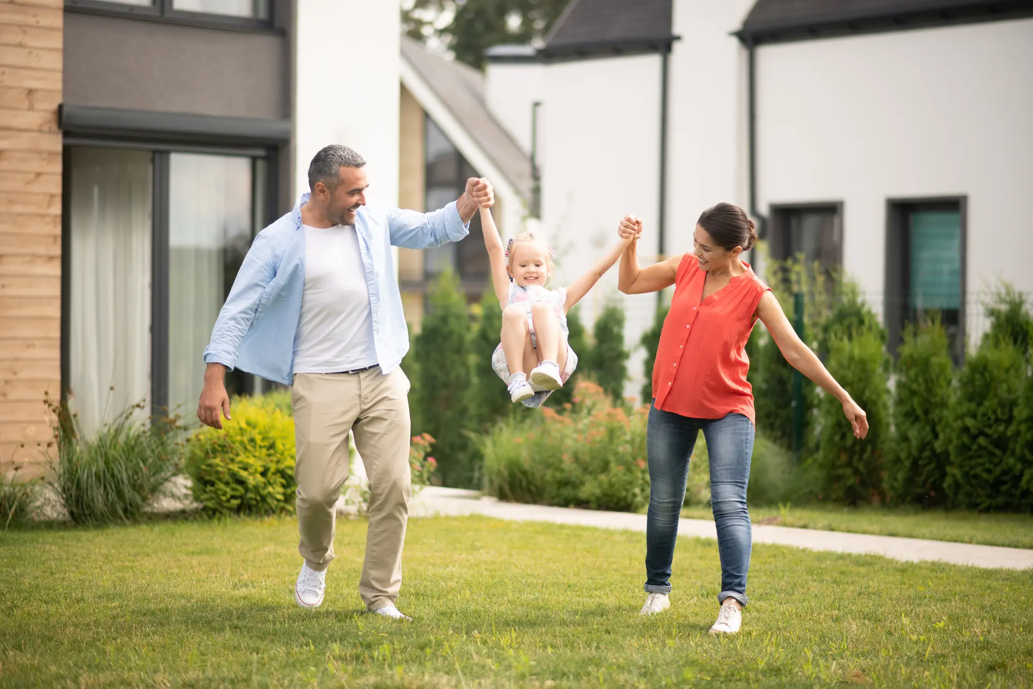 Eltern schwingen ihr Kind lachend im Garten eines modernen Hauses, grüne Hecke im Hintergrund, familiäre Szene mit Hausbezug