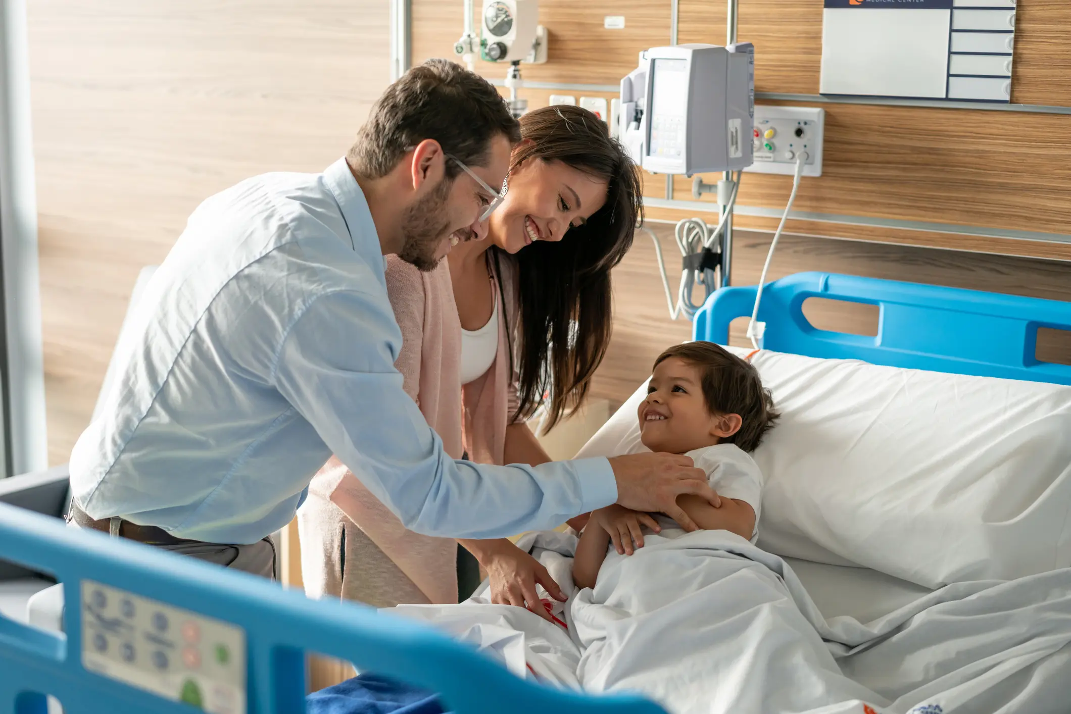 Eltern besuchen ihr lächelndes Kind im Krankenhausbett – Symbolbild für die Absicherung durch eine Zusatzversicherung