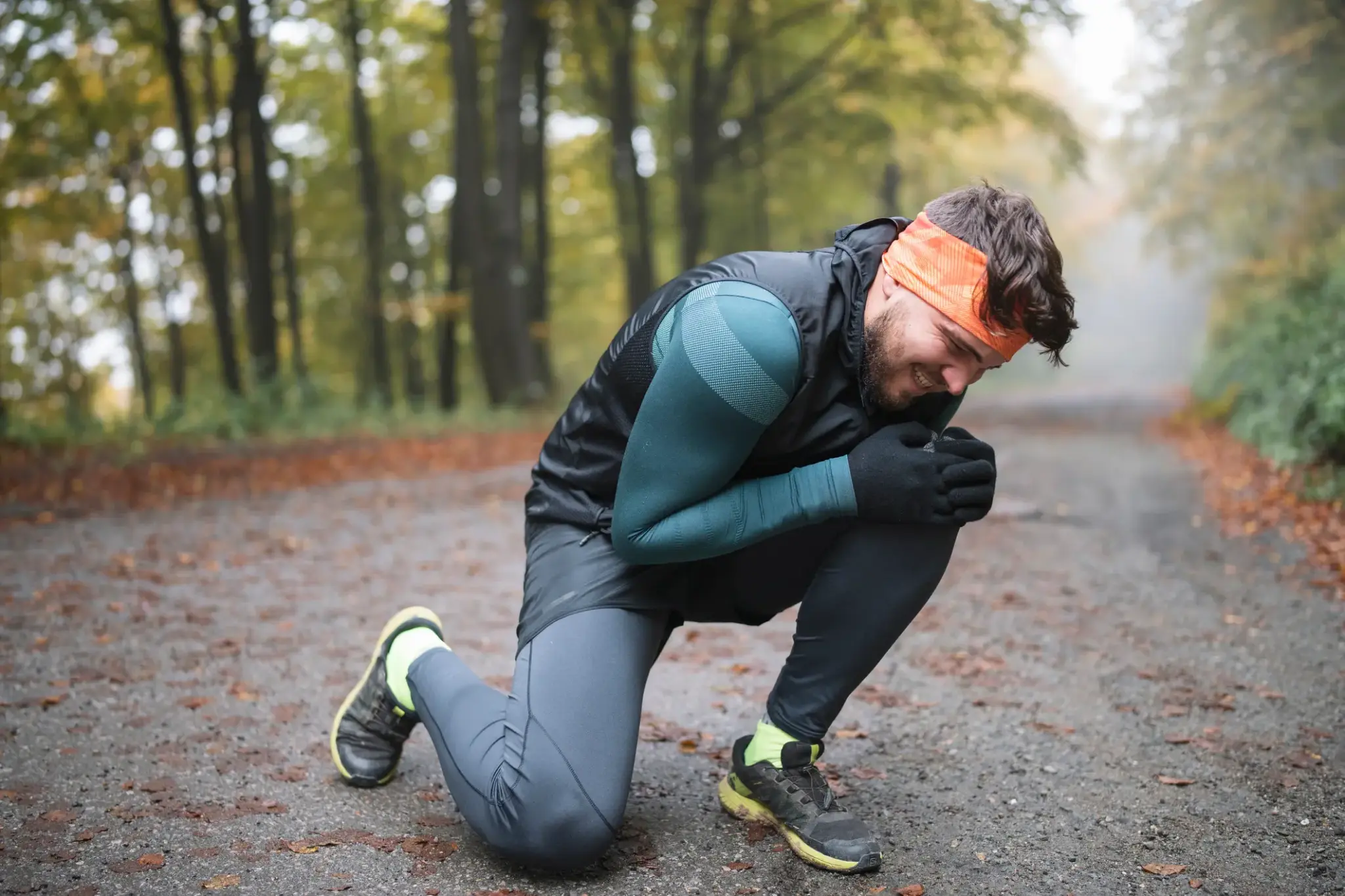 Mitwirkungsanteil | Mann kniet verletzt im Herbstwald auf dem Weg und hält sich schmerzverzerrt das Knie – Joggingkleidung und Stirnband