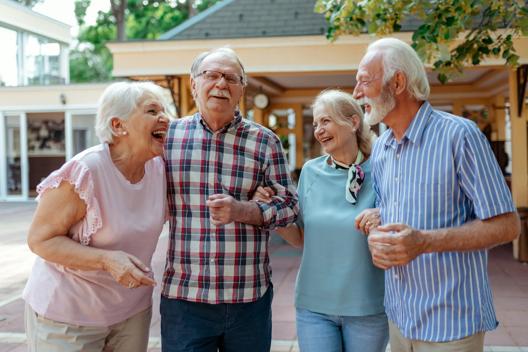 Vier ältere Menschen lachen gemeinsam im Freien und gehen Arm in Arm, gepflegte Umgebung im Hintergrund