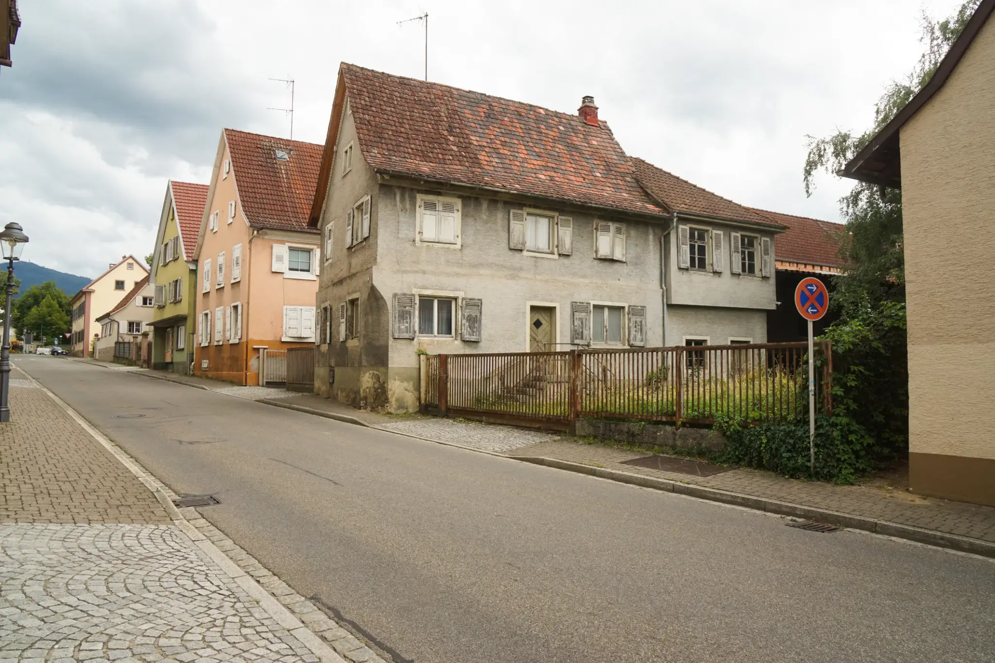 Straßenszene mit leerstehendem, sanierungsbedürftigem Wohnhaus – verwitterte Fassade, geschlossene Fensterläden, ungepflegter Garten und verlassener Eindruck.