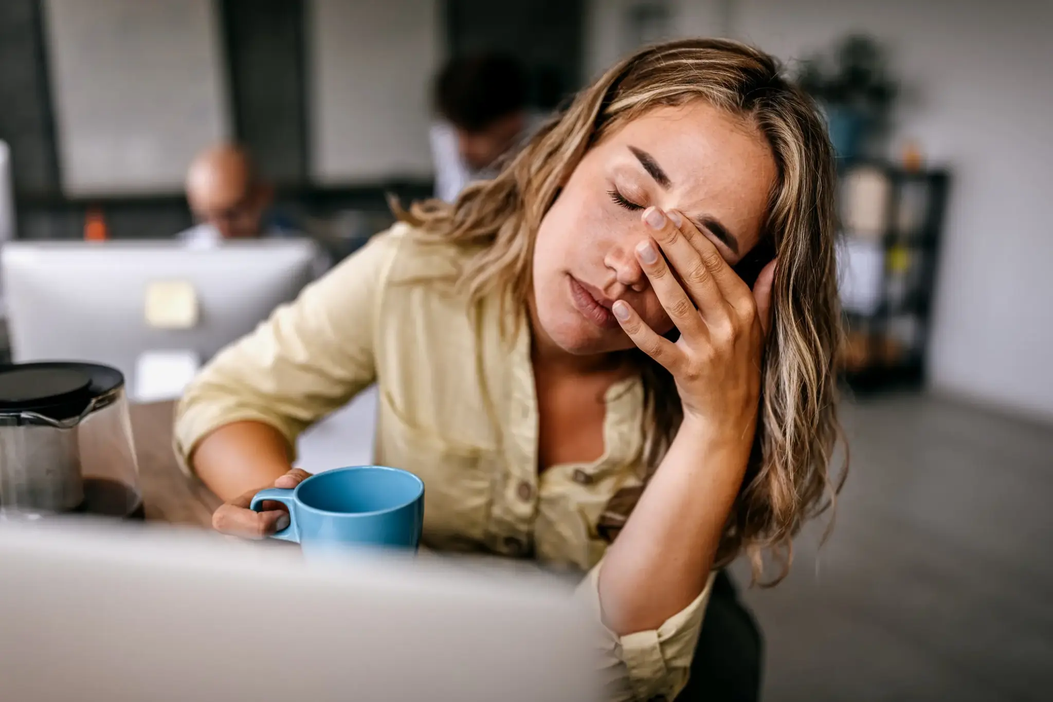 Arbeitsunfaehigkeitsversicherung | Erschöpfte Frau im Büro mit Kaffeetasse – Symbolbild für Krankheit oder Überlastung und mögliche Arbeitsunfähigkeit im Berufsalltag.