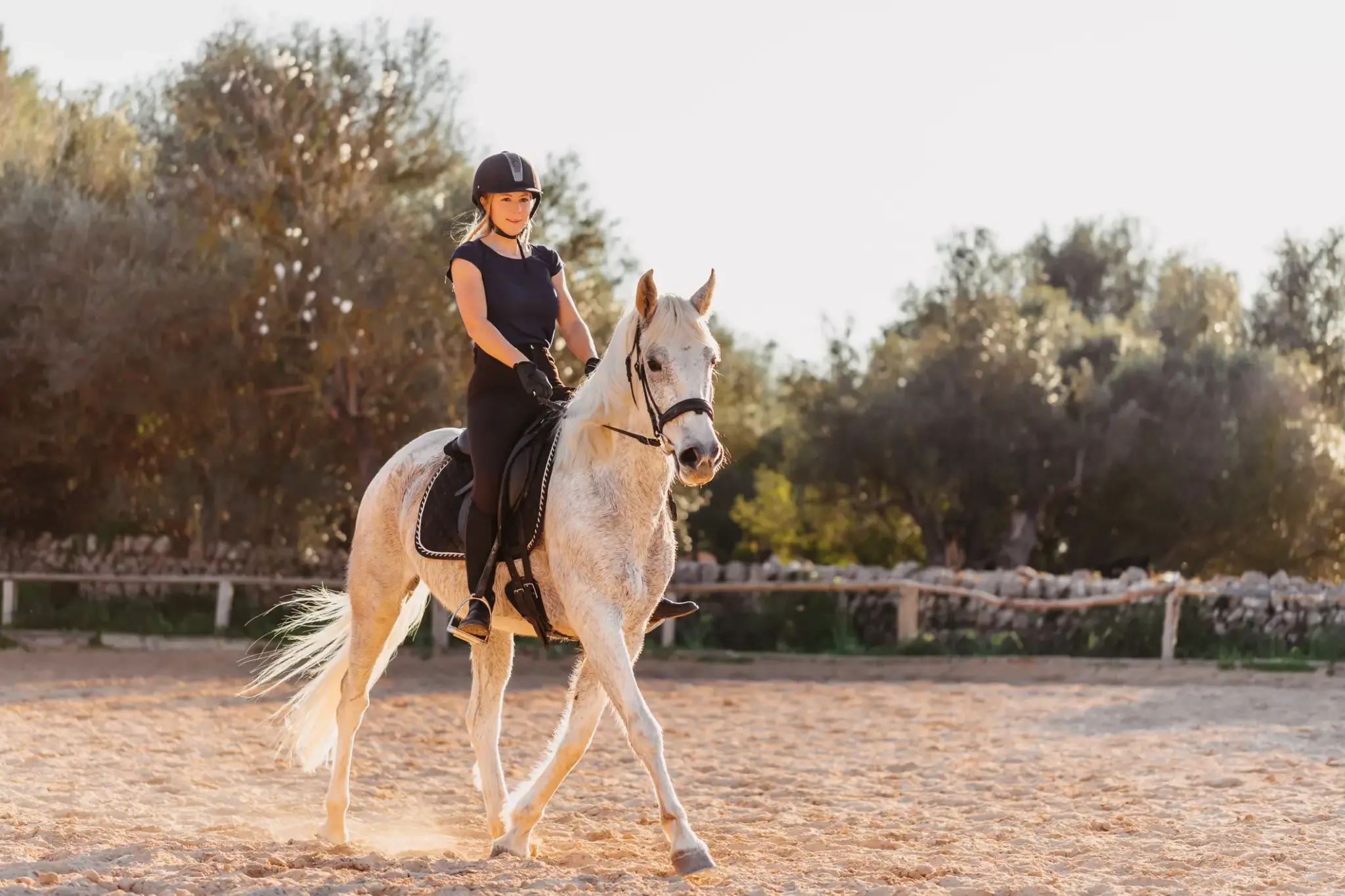 Junge Reiterin mit Helm sitzt sicher auf einem weißen Pferd auf einem Reitplatz – Symbolbild für Unfallrisiken beim Reitsport.