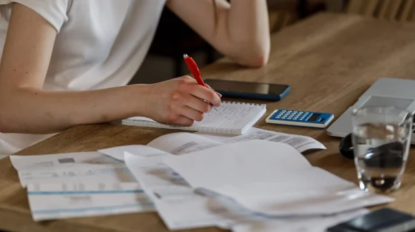 Person mit Block und Stift bei der Finanzplanung – auf dem Tisch liegen Unterlagen, Taschenrechner, Laptop und ein Glas Wasser.