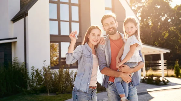 Junge Familie mit Haus im Hintergrund hält Schlüssel in der Hand – Symbolbild für Immobilie Altersvorsorge