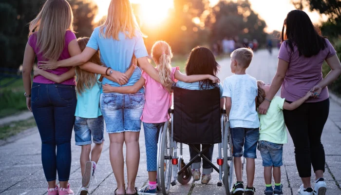 Kindergruppe von hinten, ein Kind im Rollstuhl wird liebevoll von den anderen umarmt – gemeinsamer Spaziergang bei Sonnenuntergang.