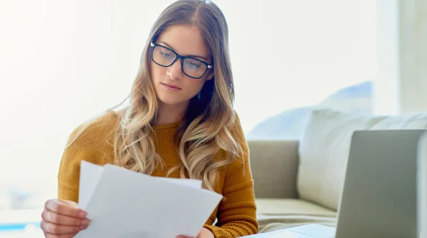 Junge Frau mit Brille prüft konzentriert Dokumente vor dem Laptop – Entscheidung zur Anpassung einer Berufsunfähigkeitsversicherung.