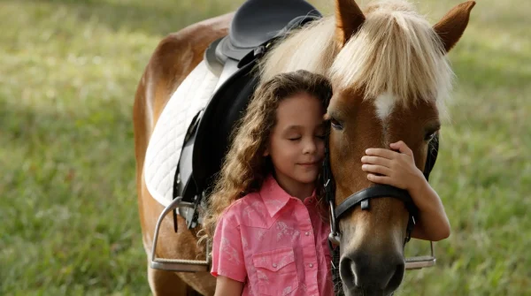 Mädchen mit rosa Hemd umarmt liebevoll ein Pony auf der Weide – Symbolbild für den Schutz durch eine Pferdeversicherung.