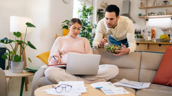 Junges Paar sitzt im Wohnzimmer auf dem Sofa, prüft Unterlagen am Laptop – Fokus auf Planung, Versicherung & Finanzen